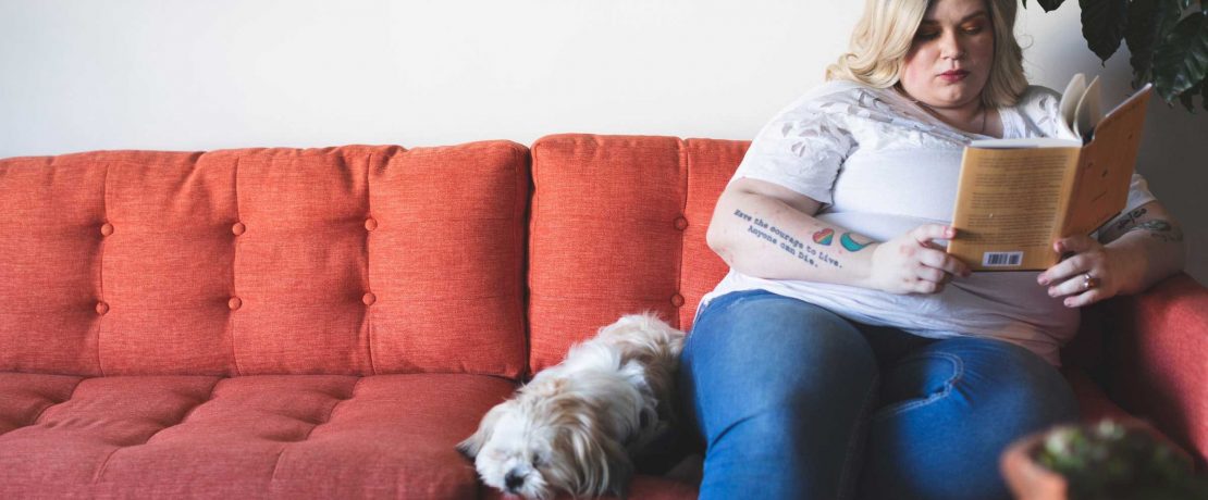 A woman sits on the end of a red sofa reading a book, with a small dog beside her