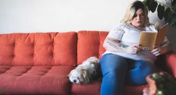 A woman sits on the end of a red sofa reading a book, with a small dog beside her