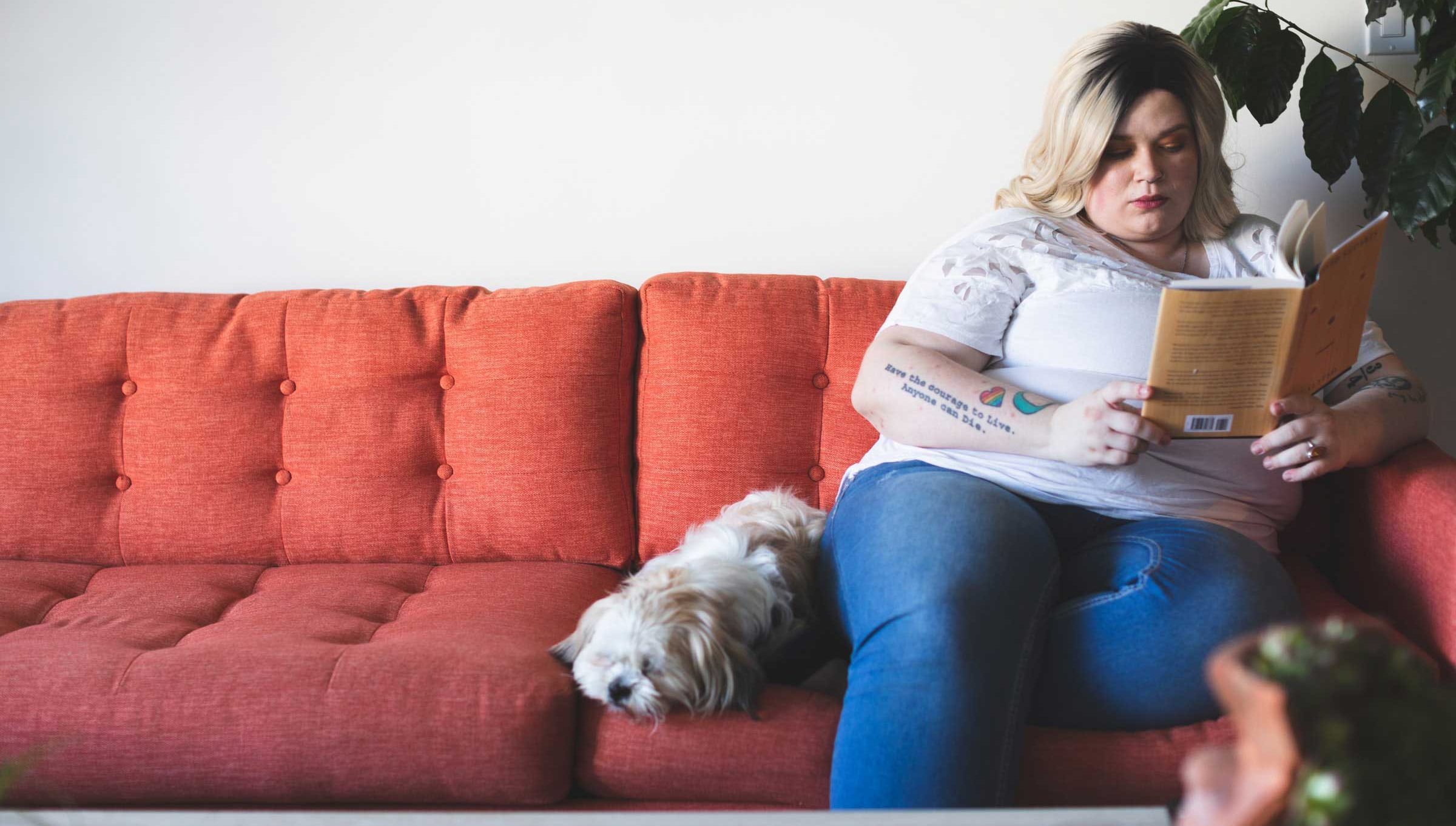 A woman sits on the end of a red sofa reading a book, with a small dog beside her