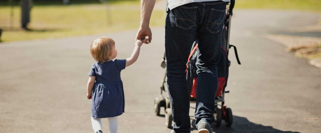 A man walks with a buggy on a path, holding a small girl by the hand