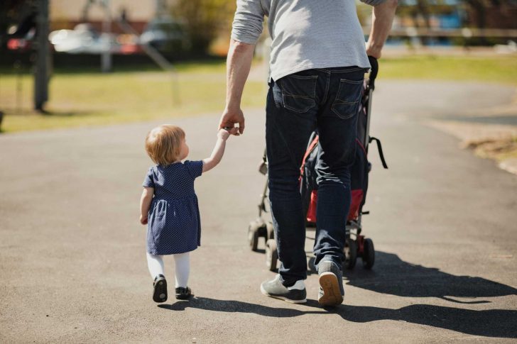 A man walks with a buggy on a path, holding a small girl by the hand