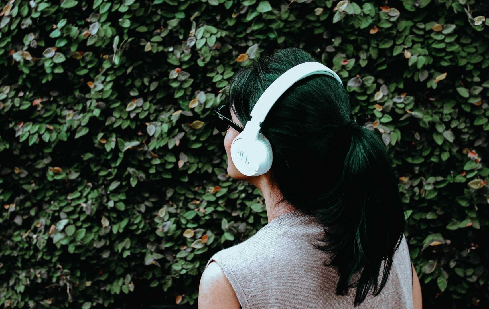 A woman wears large headphones as she faces a leafy hedge