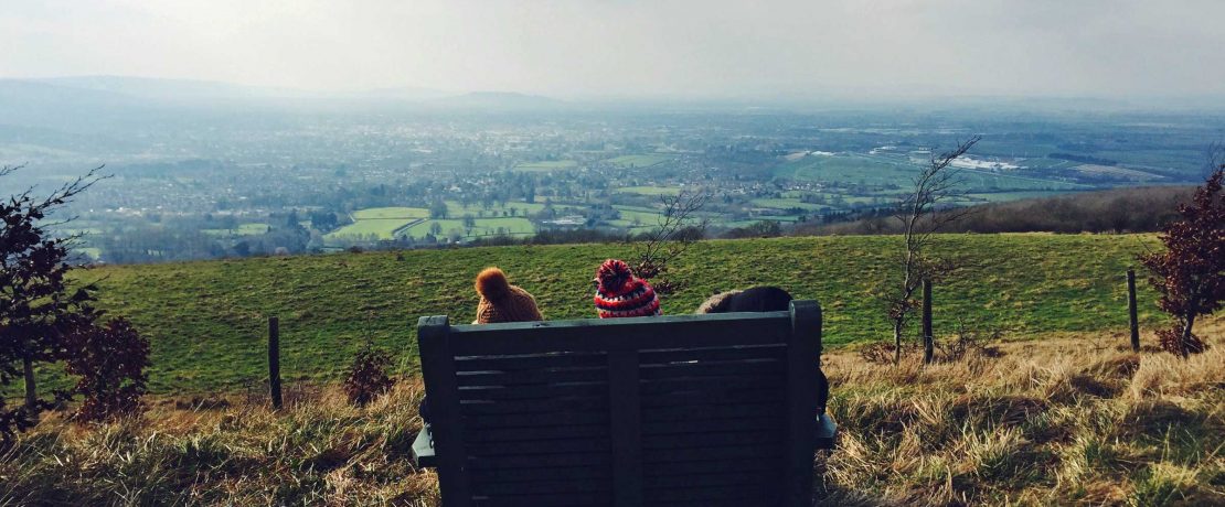 A bench is silhouetted against a countryside vista with two bobble hats visible from behind