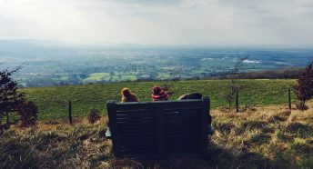A bench is silhouetted against a countryside vista with two bobble hats visible from behind