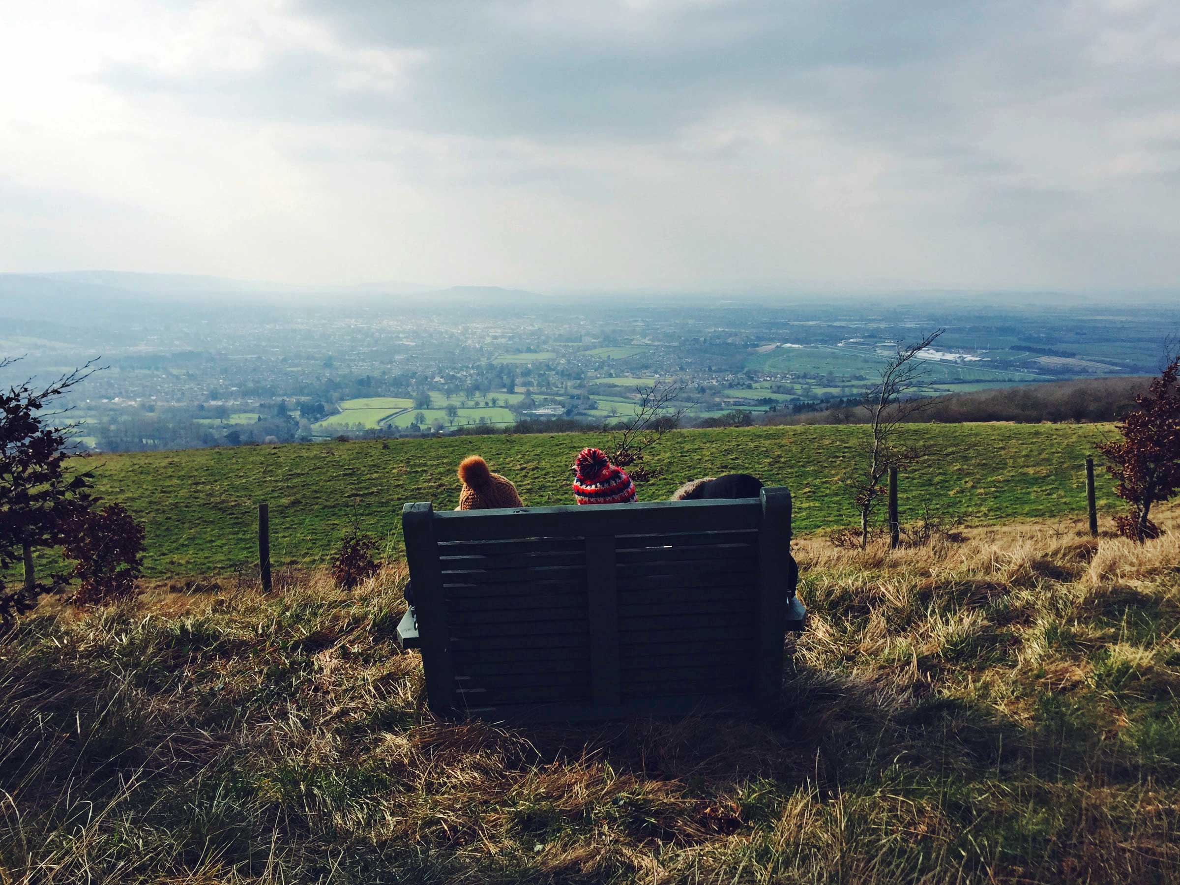 A bench is silhouetted against a countryside vista with two bobble hats visible from behind