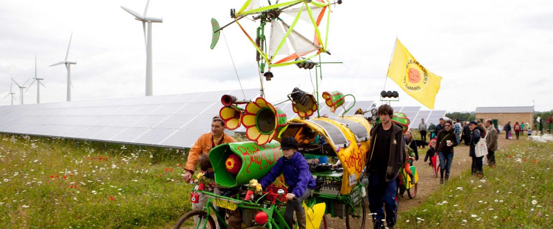 A group of people with a painted and adorned bicycle stand in front of solar panels and wind turbines