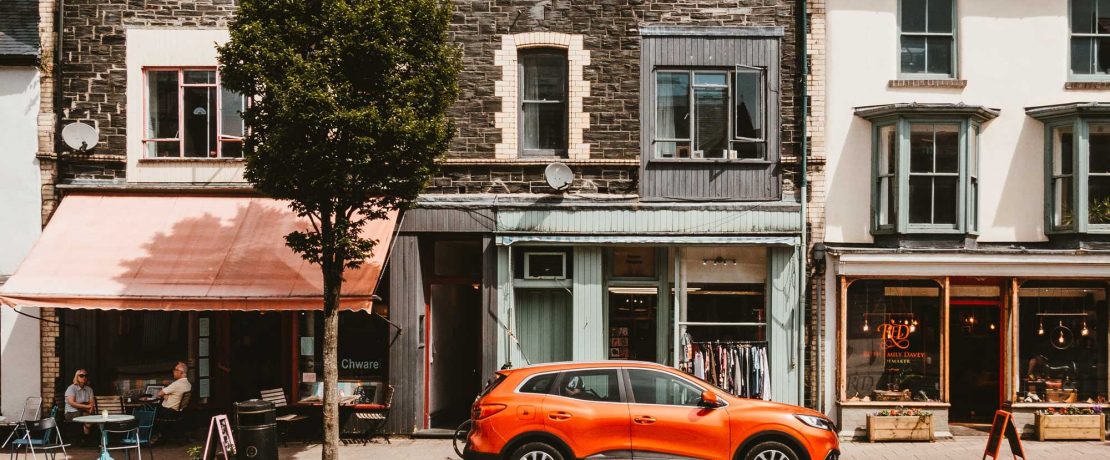 A high street with a car parked, a shop and cafe with people eating outside