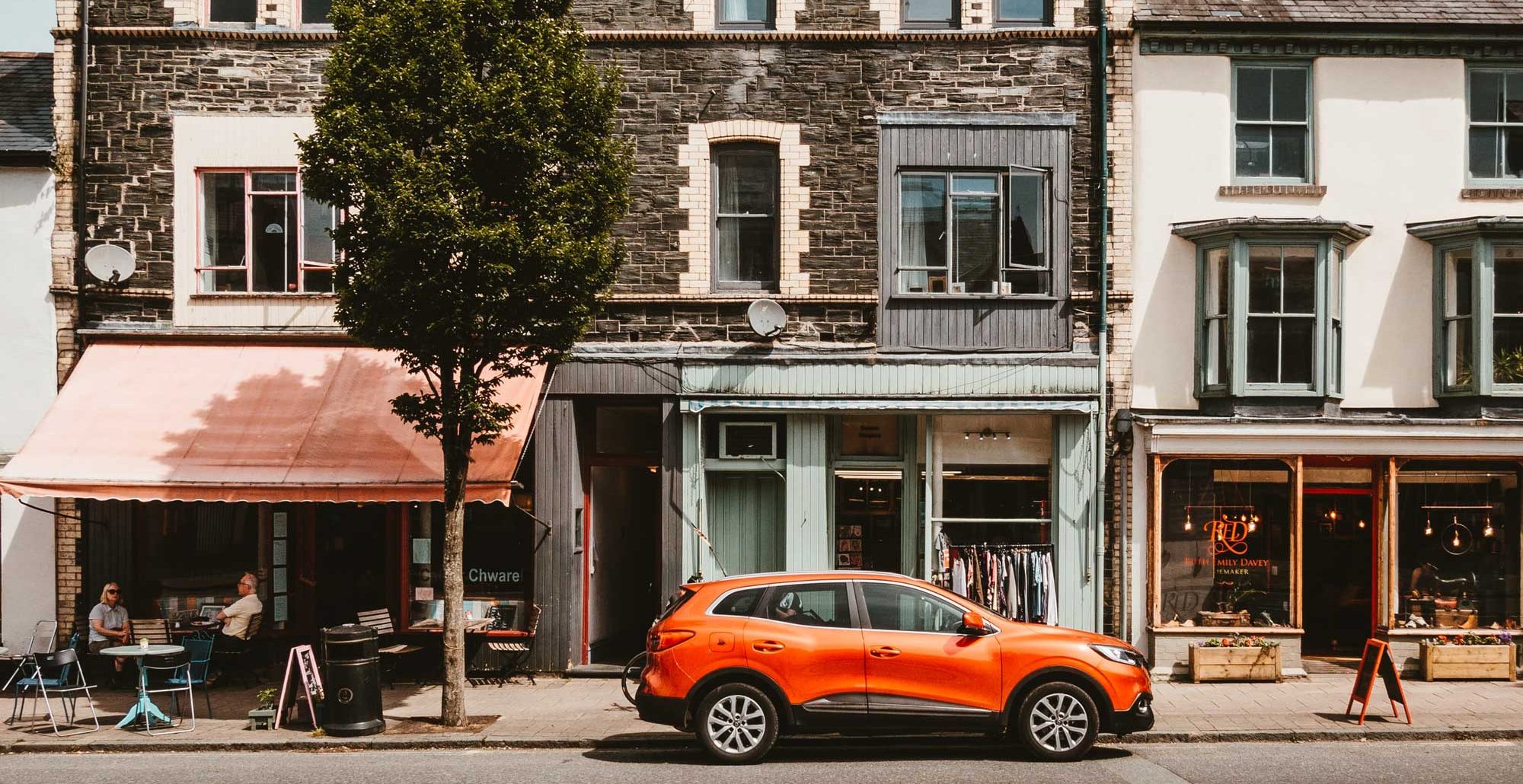 A high street with a car parked, a shop and cafe with people eating outside