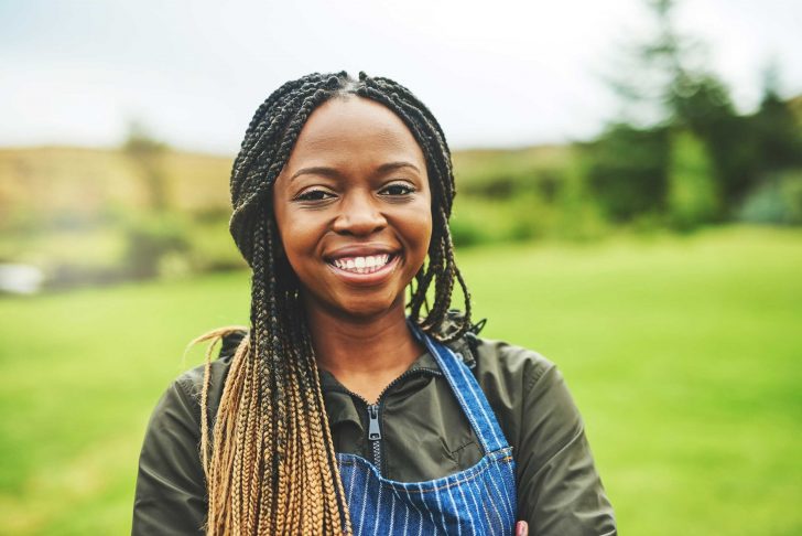 A young female farmer wearing an apron stands in front of green fields