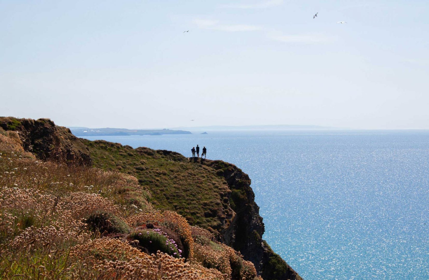 Three people stand on a striking cliff top with sea behind