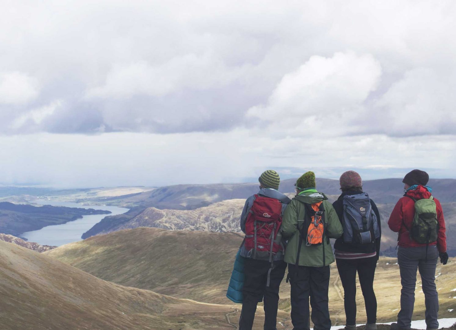 Four women in coats and hats look down on a lake from a high fell