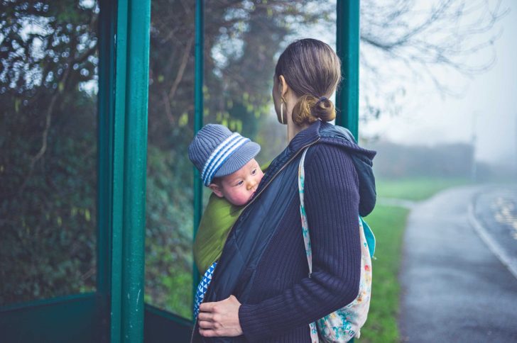 A woman stands at a rural bus stop with a baby in a sling