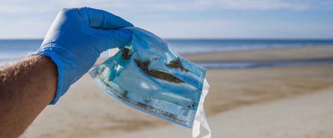 A discarded disposable face mask is held by a person on a sandy beach