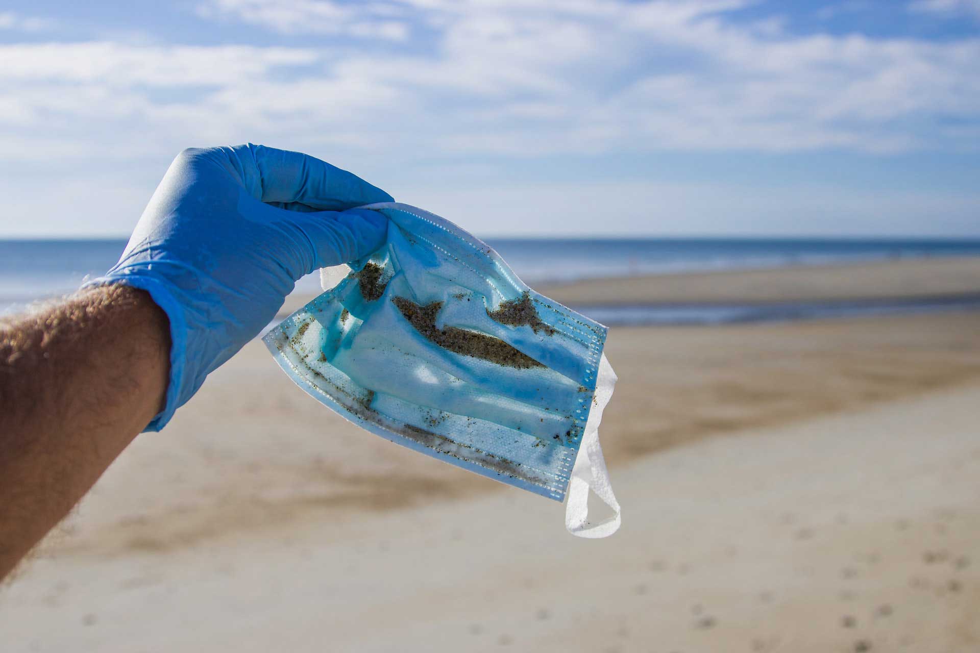 A discarded disposable face mask is held by a person on a sandy beach