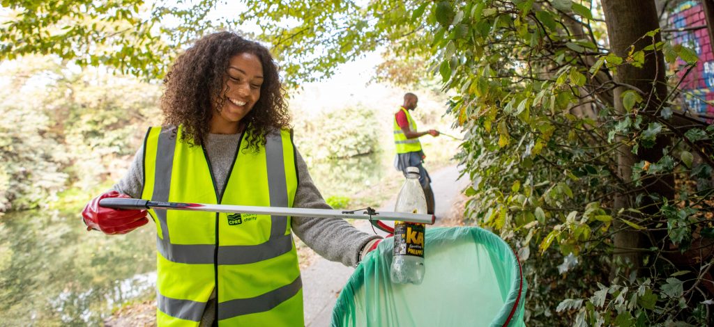 Smiling woman placing plastic bottle into recycling bag near trees
