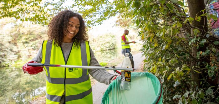 Smiling woman placing plastic bottle into recycling bag near trees