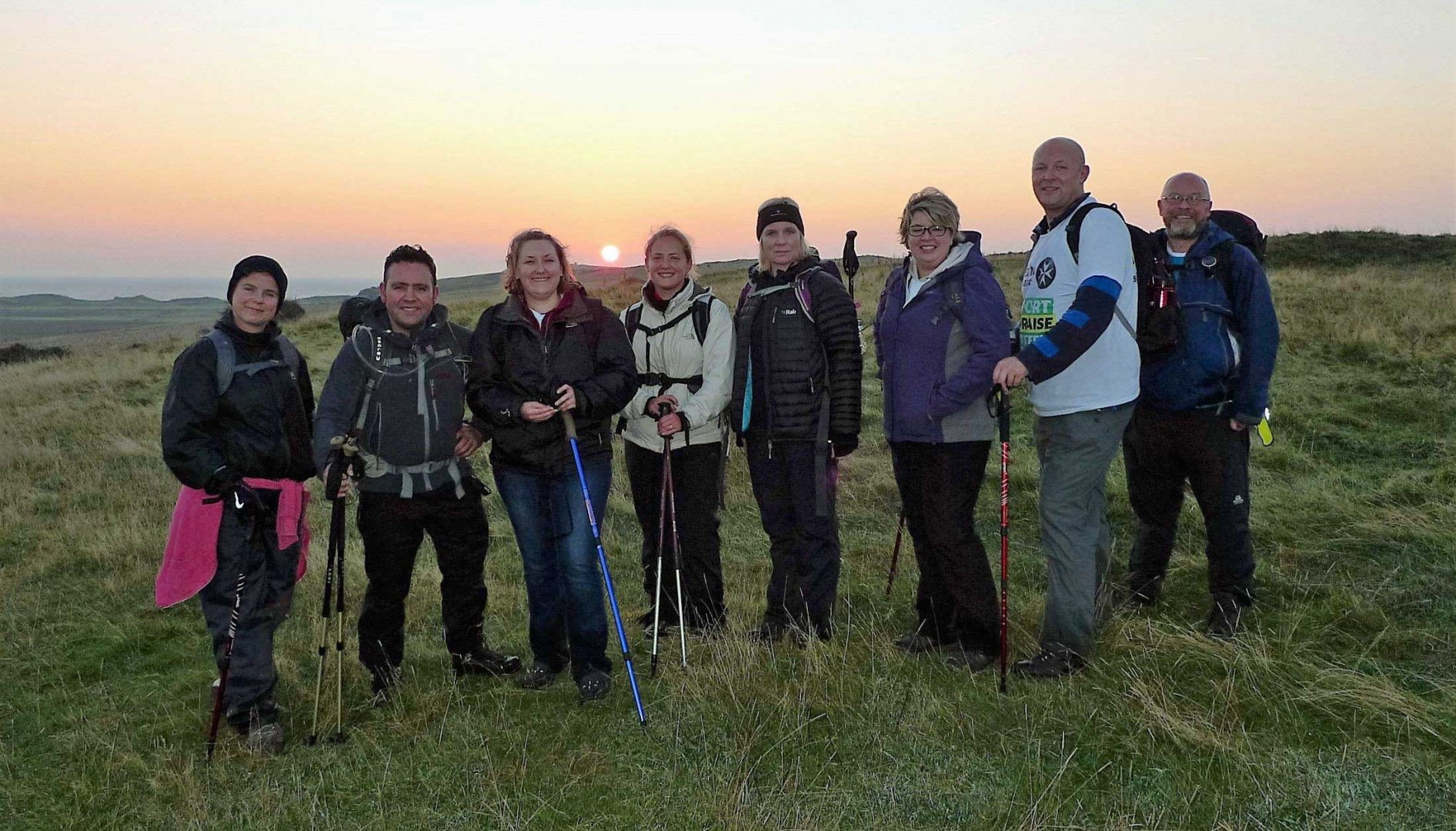 A group of walkers with sticks stand in front of a sunset