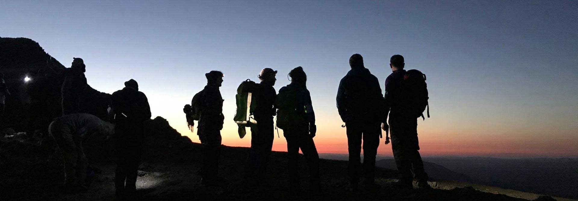A group of walkers on a hill are silhouetted against a red sky