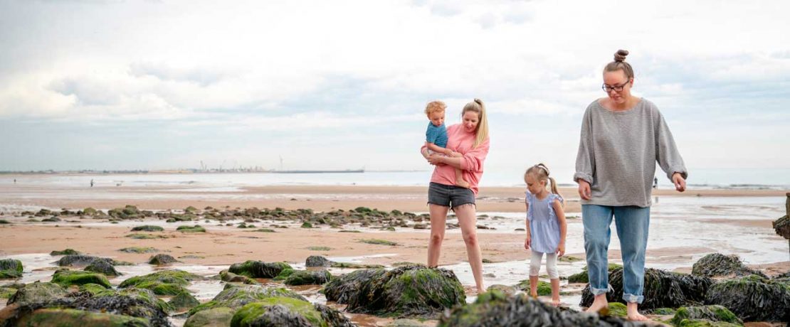 A same sex couple look down at rocks on a sandy beach with their small boy and girl