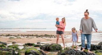 A same sex couple look down at rocks on a sandy beach with their small boy and girl