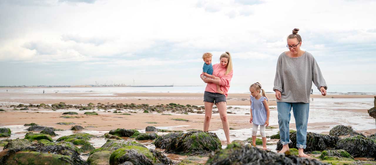 A same sex couple look down at rocks on a sandy beach with their small boy and girl