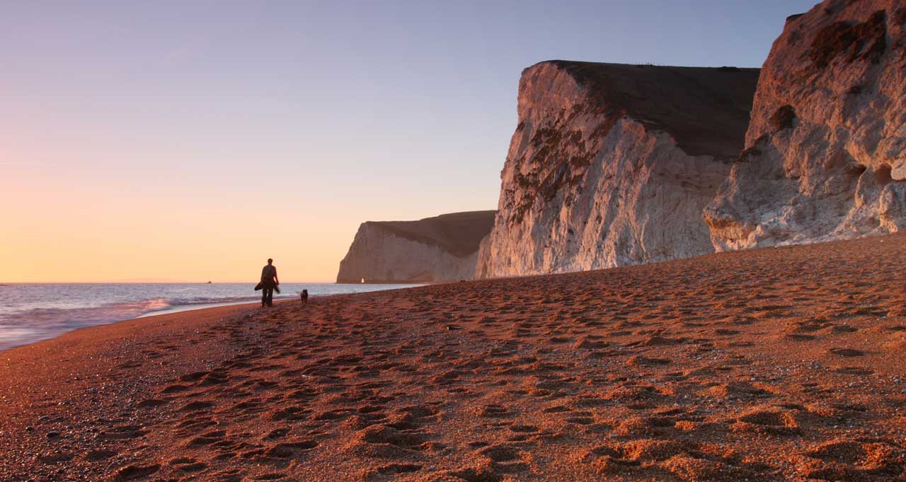 Person and dog walking on beach with sand and cliffs at sunset