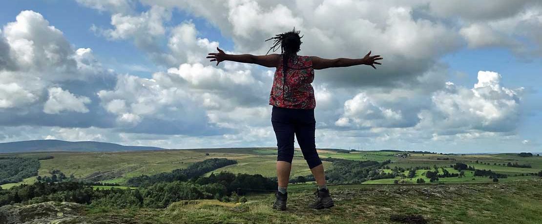 A black woman with dreadlocks faces away from the camera and stands with arms outstretched in the countryside