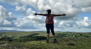 A black woman with dreadlocks faces away from the camera and stands with arms outstretched in the countryside