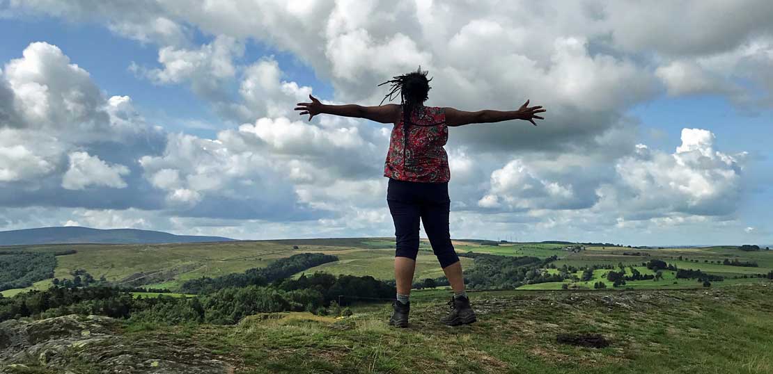 A black woman with dreadlocks faces away from the camera and stands with arms outstretched in the countryside