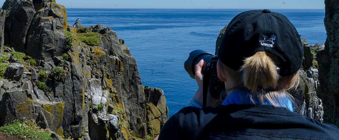 A woman with a large camera faces towards some cliffs, with her back to us