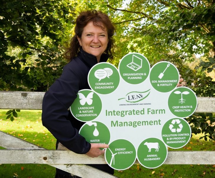 A woman in front of trees holding a display showing aims of sustainable farming