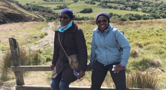 Two black women resting on a fence with a footpath, open countryside and hills behind them
