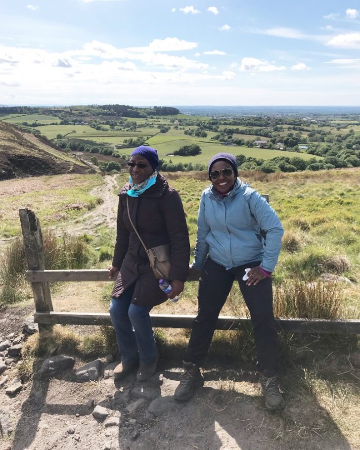 Two black women resting on a fence with a footpath, open countryside and hills behind them