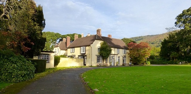 a driveway leading to a yellow house with a large lawn and green hills in the background