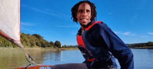 A black man in a boat on a lake with blue sky and trees in the backgrounds 