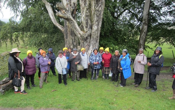 A group of older black women in coats standing in the countryside in from of a large tree trunk