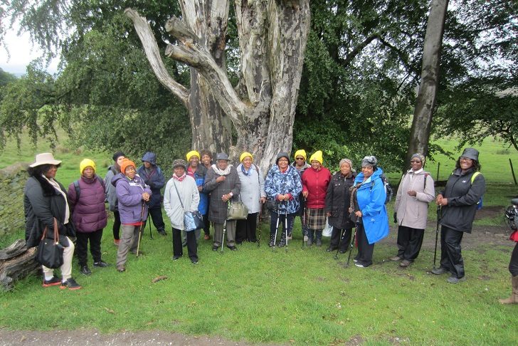 A group of older black women in coats standing in the countryside in from of a large tree trunk