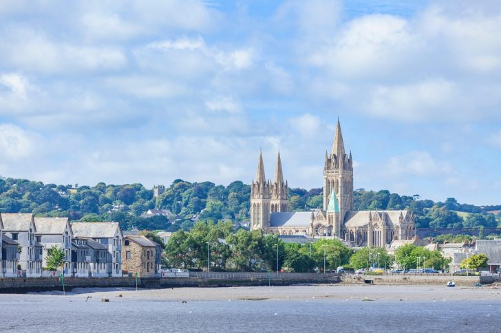 A cathedral with three spires with a river in the foreground and a tree covered hills and a parish church tower in the background