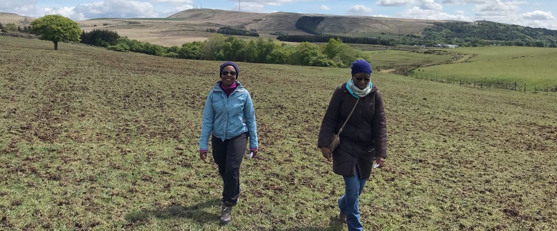 two black women walking on flat grassland with hills and blue, cloudy skies behind them