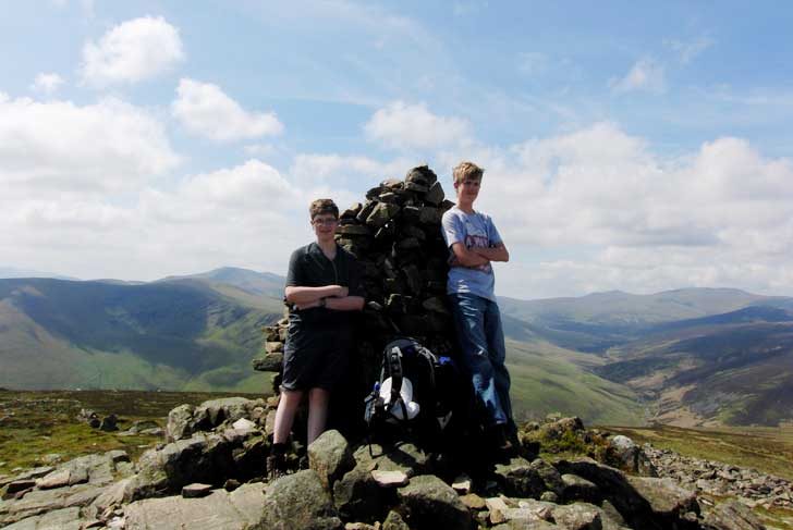 Two teenage boys lean against a trig point at the top of a fell