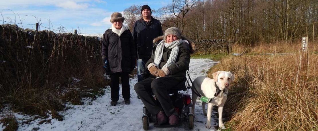 Three people and a dog on a snowy path; one is using a wheelchair