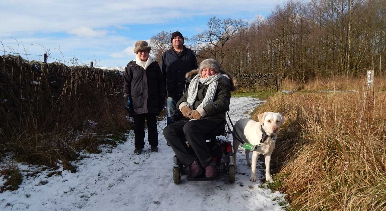 Three people and a dog on a snowy path; one is using a wheelchair