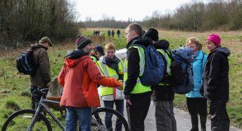 A man in a hi-vis vest and rucksack greeting a man in a red jacket with a cycle on a footpath in green space among a group of people