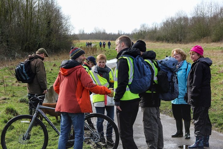 A man in a hi-vis vest and rucksack greeting a man in a red jacket with a cycle on a footpath in green space among a group of people