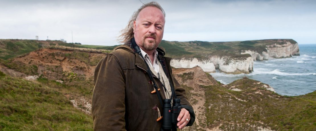 A main in a duffel coat holding binoculars in front of a rocky coastline