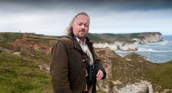 A main in a duffel coat holding binoculars in front of a rocky coastline