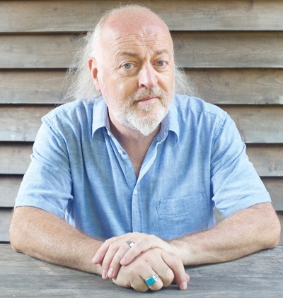 A man with white hair and beard sitting in front of a wooden fence