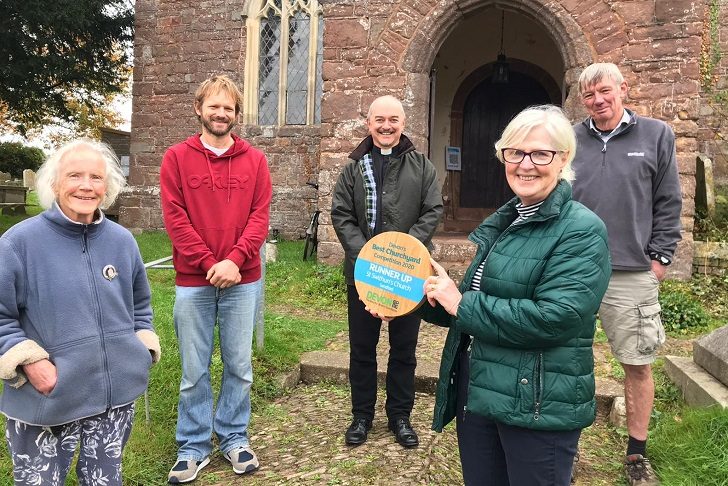 a group of smiling people standing outside the entrance of a parish church with one person holding a circular wooden plaque with a blue stripe.