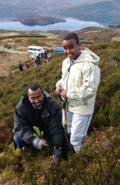 A black man and boy planting a tree on a hillside with a lake in the background