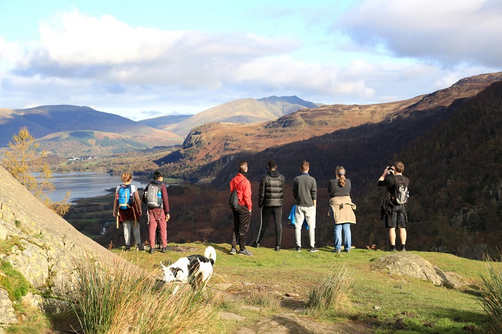 A mixed group of young people on the top of a hill looking over a valley with lakes and mountains in the distance